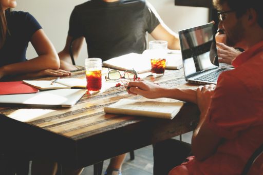 A group of people sitting around a wooden table with notebooks, glasses of iced tea, and a laptop.